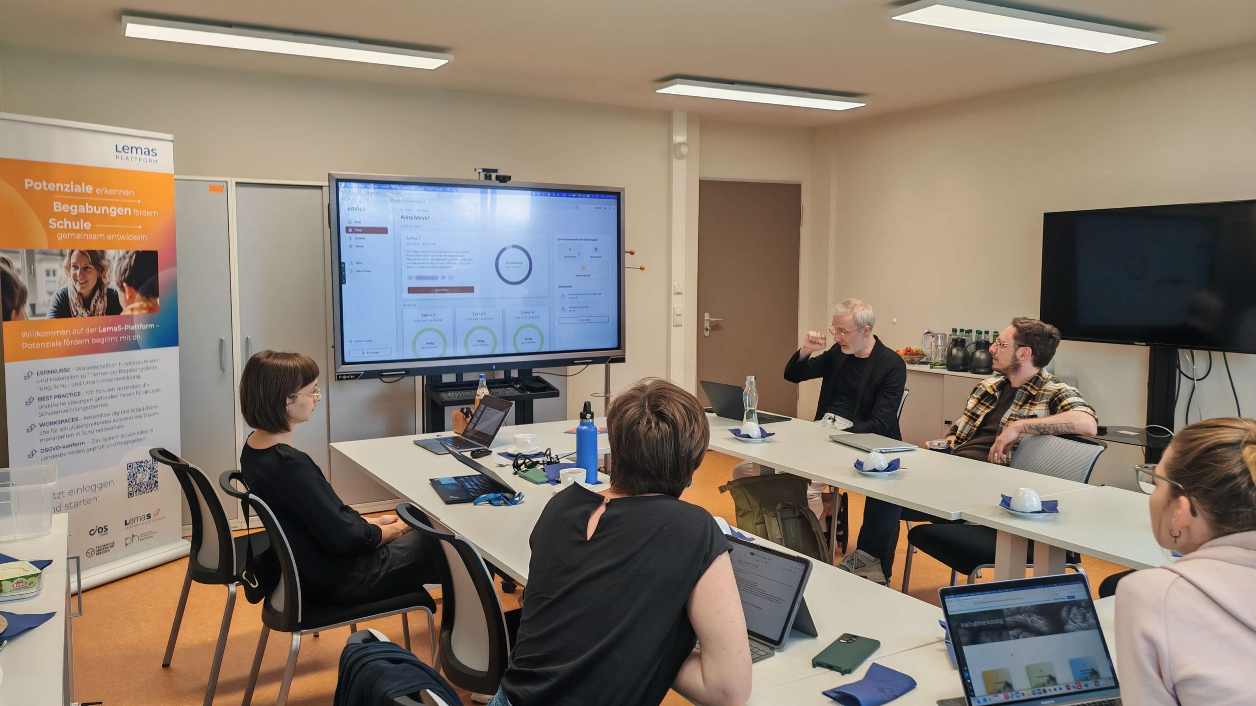 Personen am Besprechungstisch blicken auf einen Bildschirm mit der Förderplanungs-Software; daneben ein Roll-up der LemaS-Plattform.